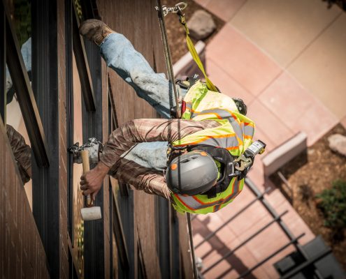 A worker uses a hammer while hanging from a rope off a building