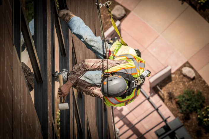A worker uses a hammer while hanging from a rope off a building